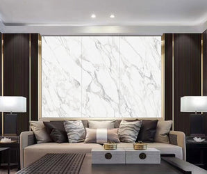 A modern living room featuring a beige sofa, multiple cushions, two table lamps, elegant decor, warm lighting, and a luxurious marble wall adorned with Calacatta White.