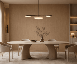 A modern dining room featuring the “Natural Ash” oval table, complemented by four beige chairs, a minimalist pendant light, a vase with branches, neutral walls adorned with a decorative wall panel, and built-in shelves displaying curated decor items. Natural light floods the space through a large window.