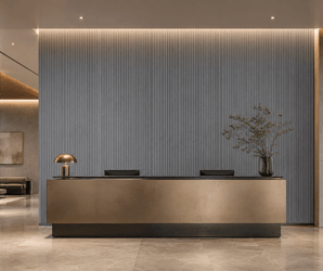 Modern reception desk with a gold front, black countertop, desk lamp, and vase with green branches. The background features the Silver Ash Fluted PS Wall Panel in gray, adding texture to the spacious, elegant lobby.